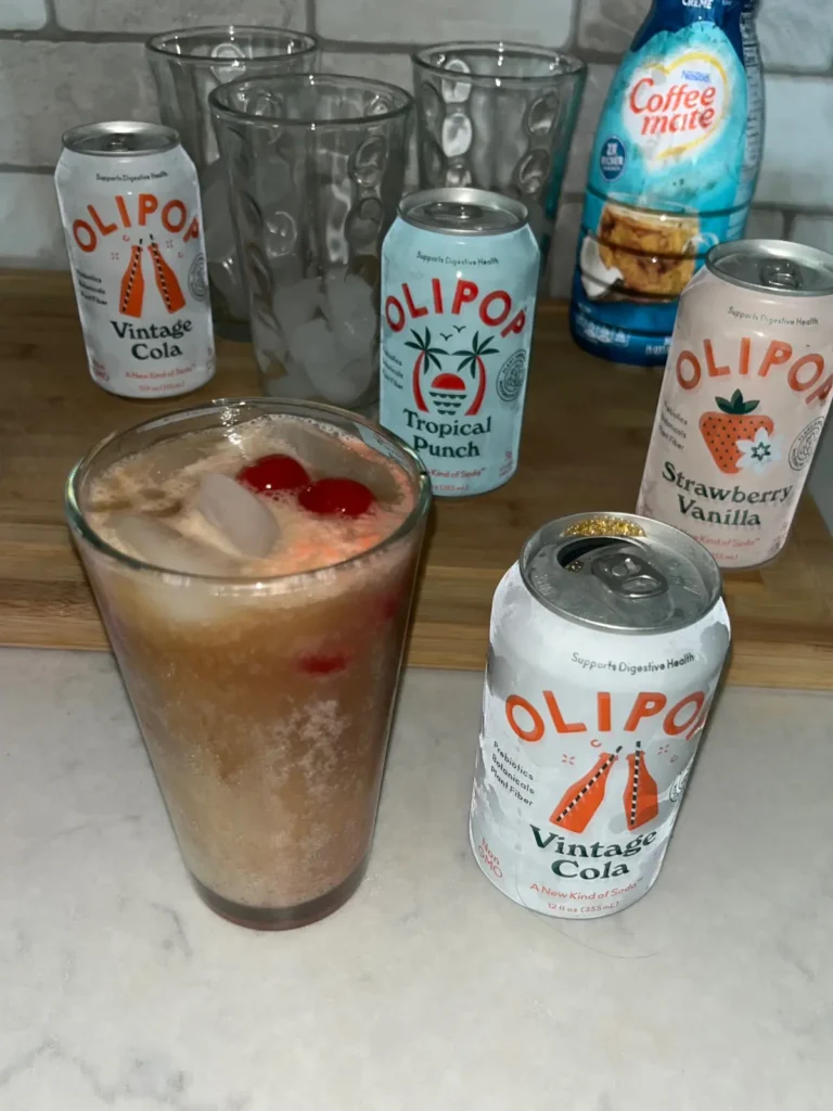 A cherries and cream dirty soda in a large glass on the counter next to a can of Olipop with more cans in the background.