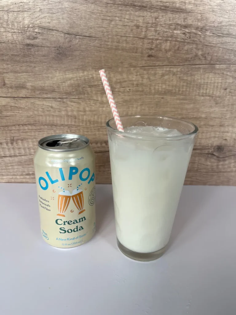 A dirty soda with Olipop cream soda flavored in a large glass with a straw next to a can of Olipop.