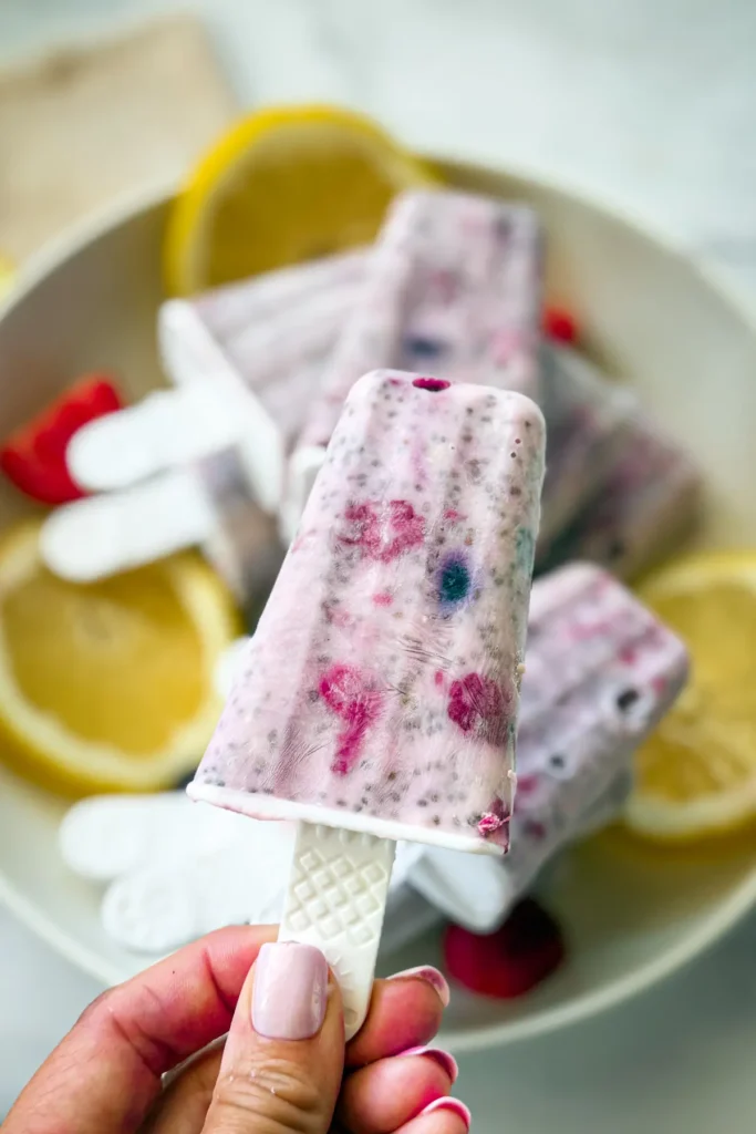 Someone holding a lemon blueberry and raspberry popsicle up with a plate of them in the background. 