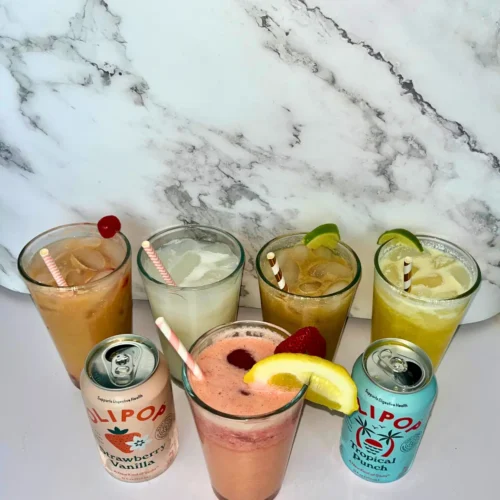5 dirty sodas with Olipop in a large clear glass with straws in it on a counter next to cans of Olipop.