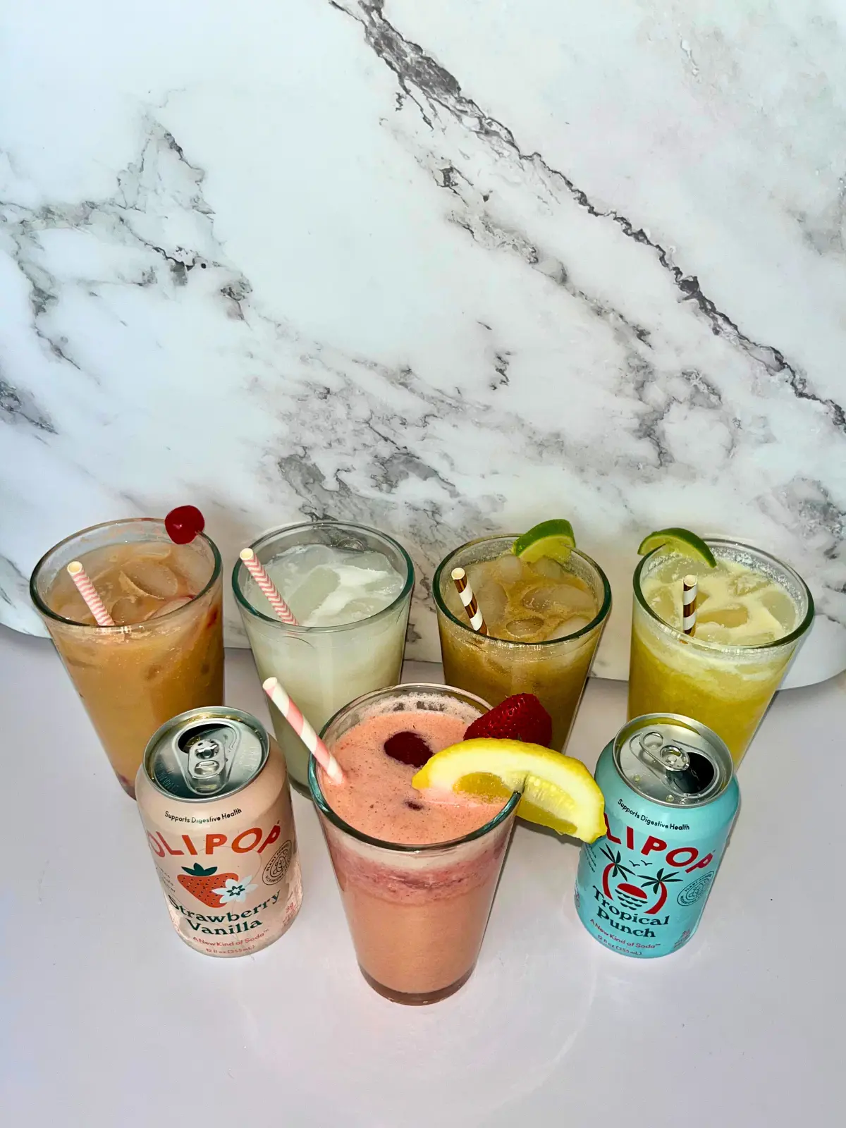 5 dirty sodas with Olipop in a large clear glass with straws in it on a counter next to cans of Olipop.