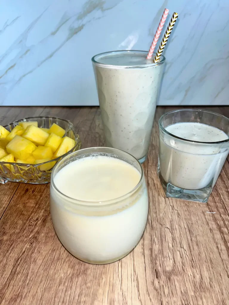 Pineapple kombucha smoothies with protein powder in glasses on a counter next to a bowl of fresh pineapple.