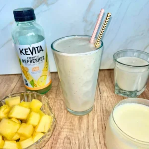 A large glass of pineapple protein kombucha smoothie next to smaller glasses and a bottle of KeVita kombucha.