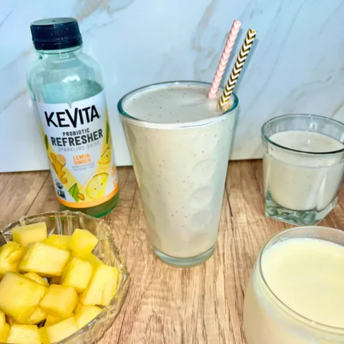 A large glass of pineapple protein kombucha smoothie next to smaller glasses and a bottle of KeVita kombucha.