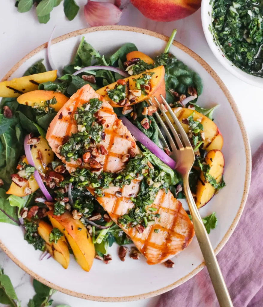 A superfood salmon salad with chimichurri and grilled peaches in a white bowl with a fork on a table.