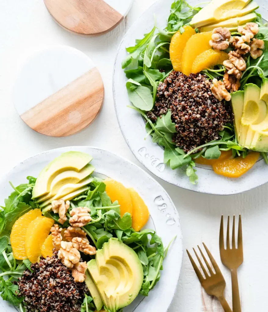 Two arugula salads topped with oranges, chopped walnuts, avocado and quinoa on a table.