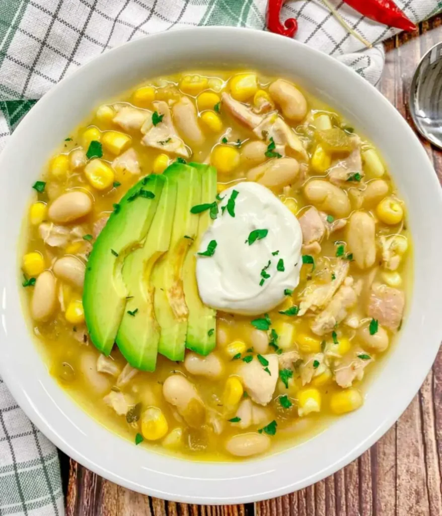 A bowl of white chicken chili topped with sliced avocado and sour cream on a table.
