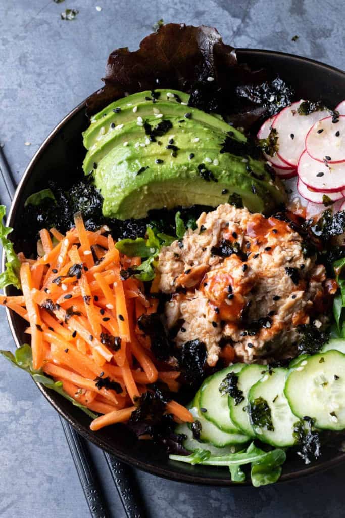 Spicy salmon salad with sliced avocados and diced carrots in a black bowl.