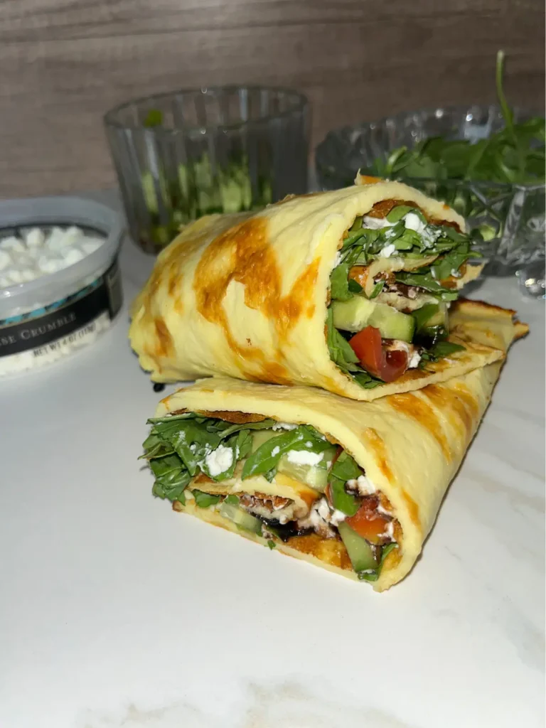 Two cottage cheese flatbreads stuffed with arugula salad on a cutting board with a bowl of goat cheese, tomatoes and cucumbers in the background.