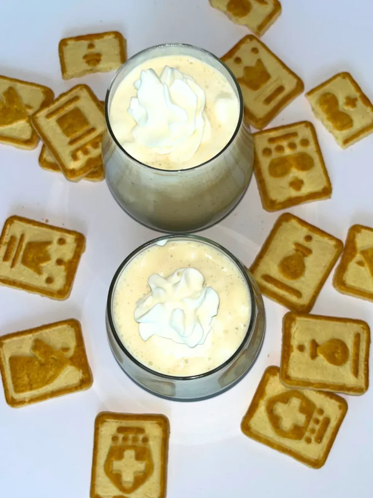 Two banana pudding protein shakes in small glasses, topped with whipped cream and surrounded by Chessmen cookies on a counter.