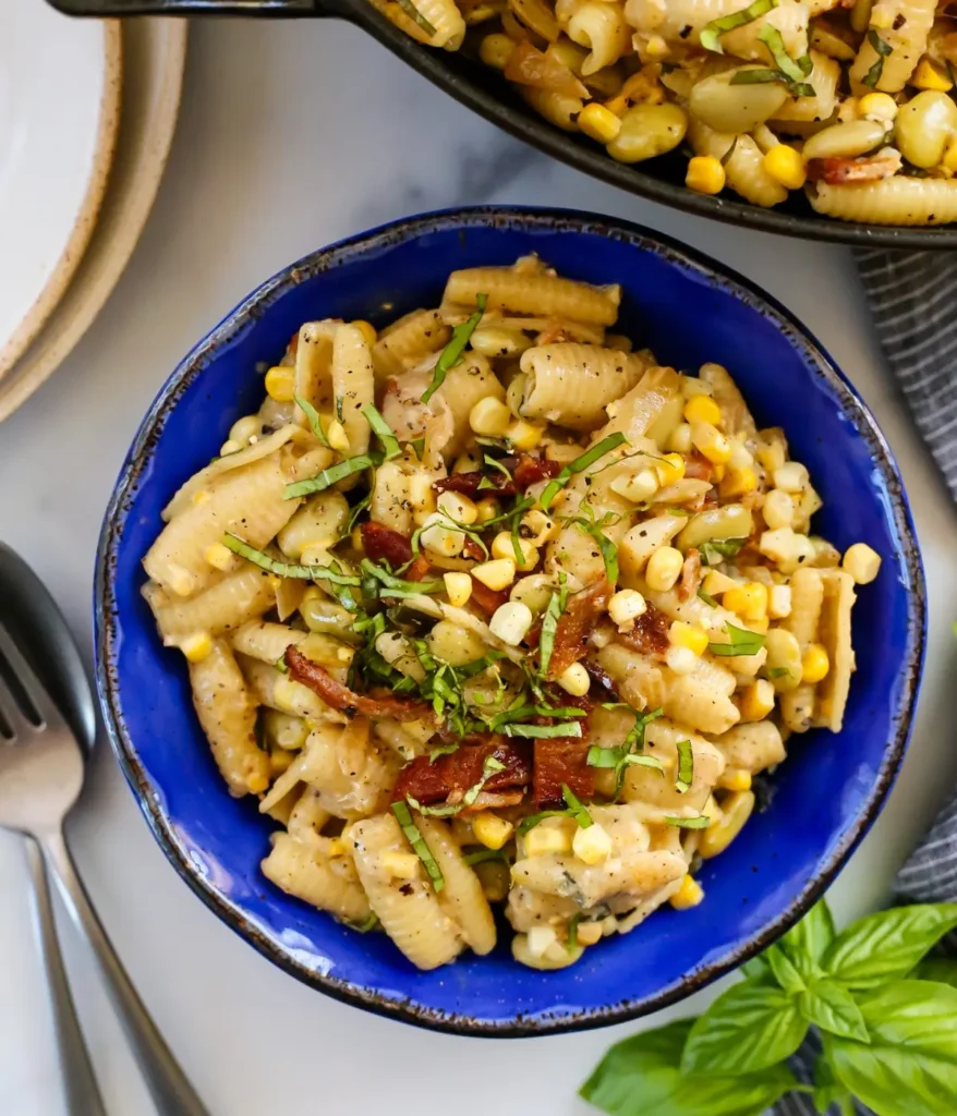 Butter bean and corn pasta in a blue bowl on a table next to a large platter of pasta.