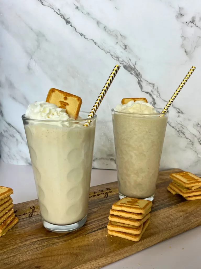 Two larges glasses filled with the banana pudding protein shake, topped with whipped cream, a Chessmen cookie and a straw.