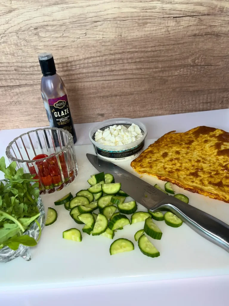 A cottage cheese flatbread, diced cucumbers, a bowl of tomatoes, balsamic glaze, and arugula on a cutting board with a knife.