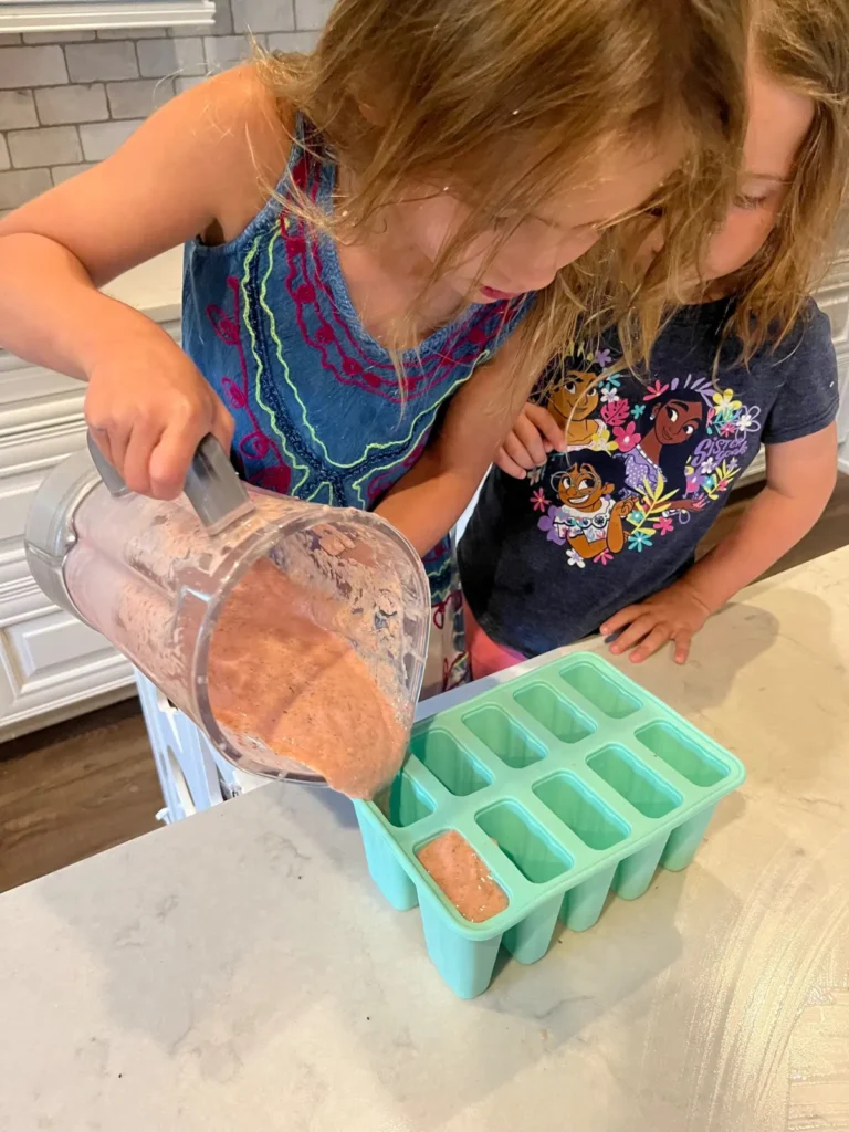 Two young girls assisting with making popsicles by pouring blender ingredients into popsicle molds.