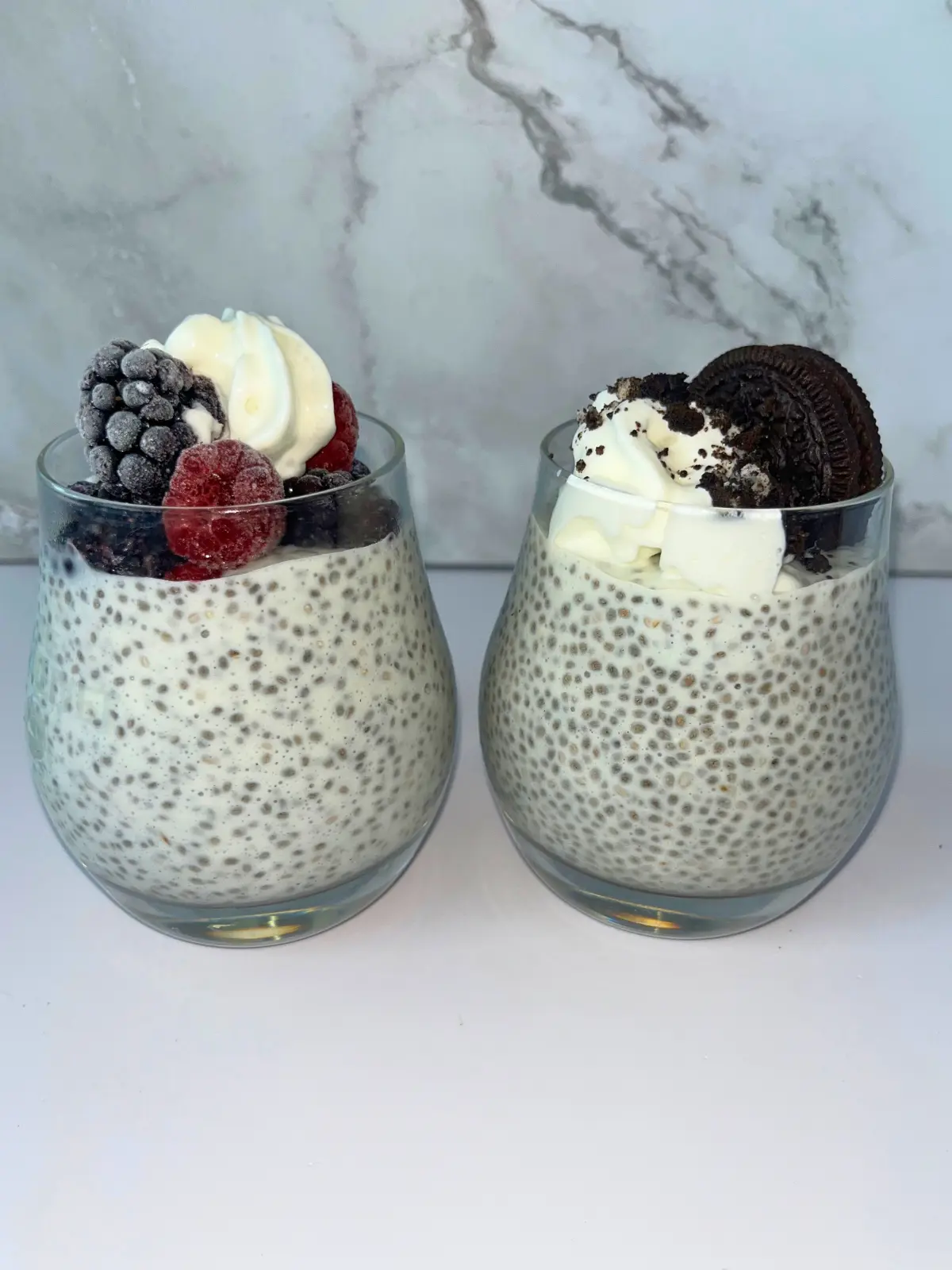 Two kefir chia puddings in glass cups, one topped with berries and whipped cream and the other with Oreos and whipped cream.