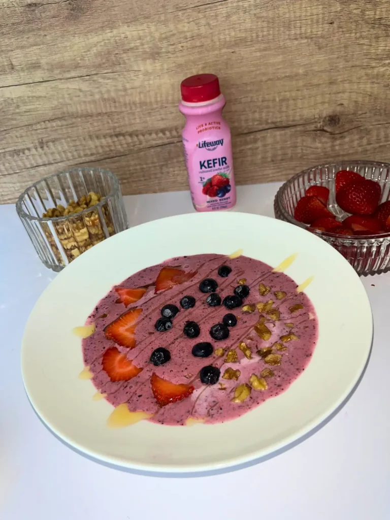 A fully assembled smoothie bowl next to a bottle of Lifeway kefir, a bowl of strawberries, and a bowl of chopped walnuts on a counter.