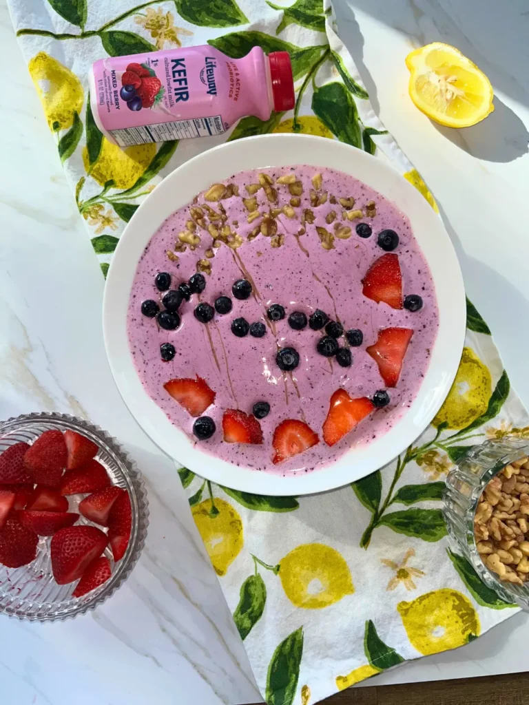 A lemon berry smoothie bowl with kefir on a colorful lemon towel next to a bowl of strawberries and a bottle of kefir on a counter.