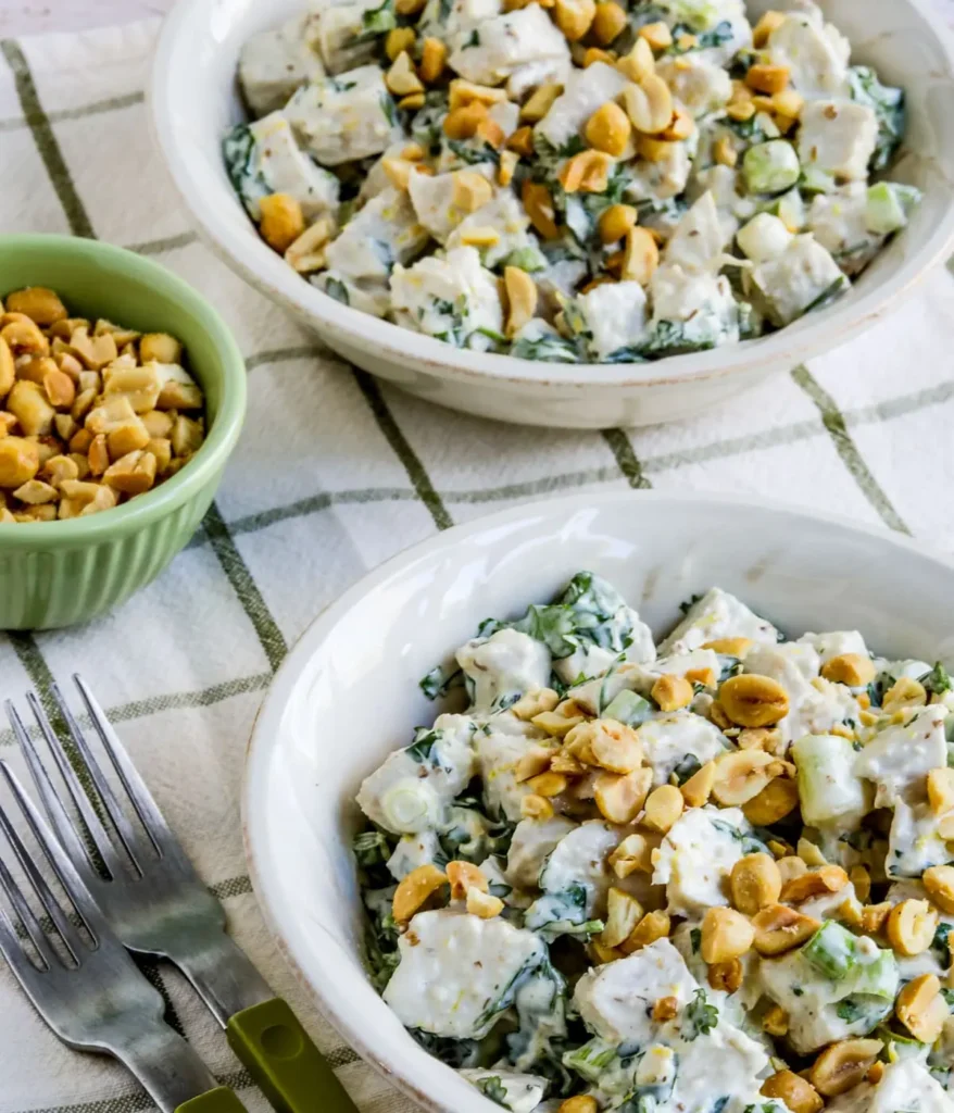 Two bowls of spicy chicken salad on a table next to a bowl chopped nuts.