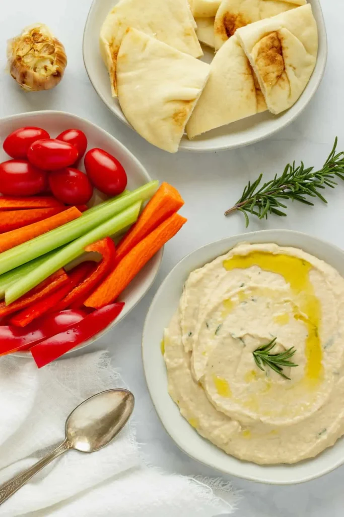 A bowl of white bean dip on a table next to a bowl of pita wedges and diced vegetables for dipping.