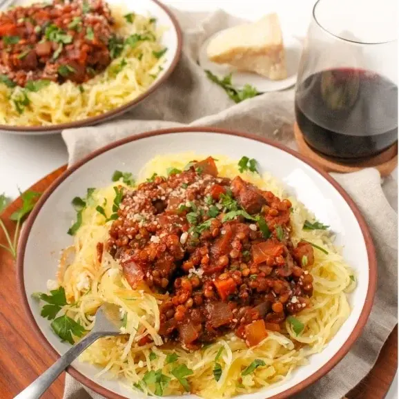 A bowl of instant pot lentil and spaghetti squash bolognese with a fork in it on a table with a glass of wine.