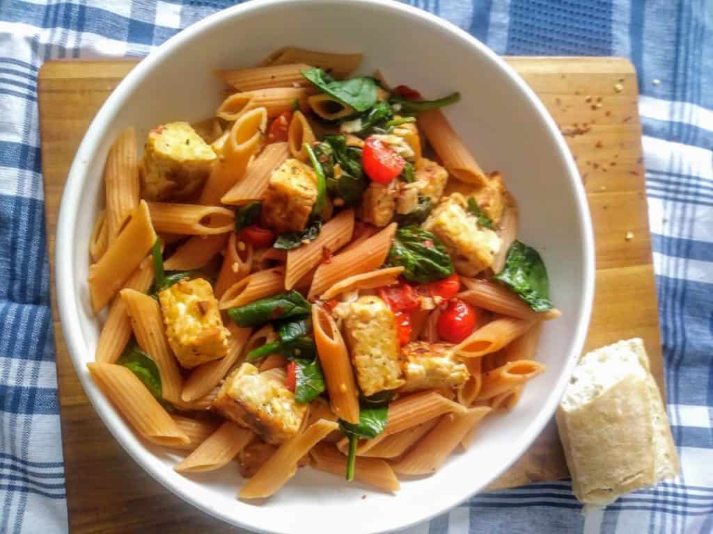 Lentil pasta with tempeh and cooked veggies in a bowl on a wood cutting board.