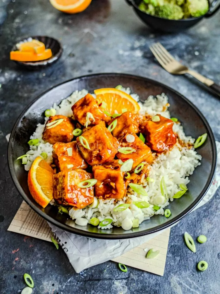 Crispy tofu with a sticky orange sauce over a bed of rice in a bowl on a table.