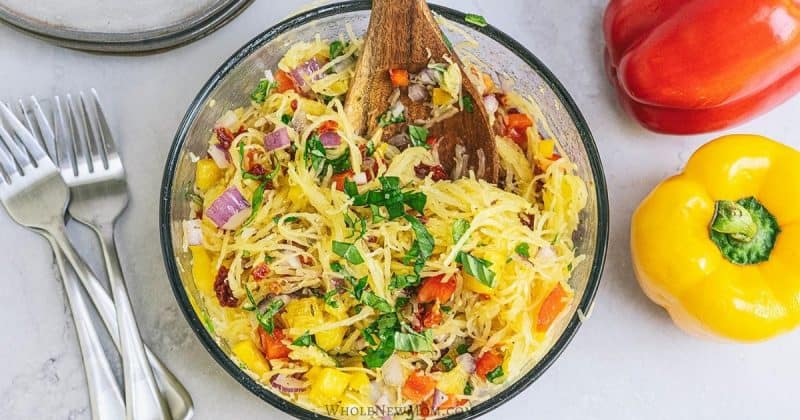 A spaghetti squash salad with veggies in a large glass bowl on a table next to silverware and bell peppers.