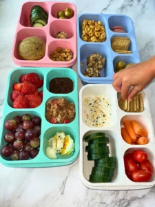 4 snack boxes of various colors filled with different snacks on a counter with someone reaching into one to grab a snack.