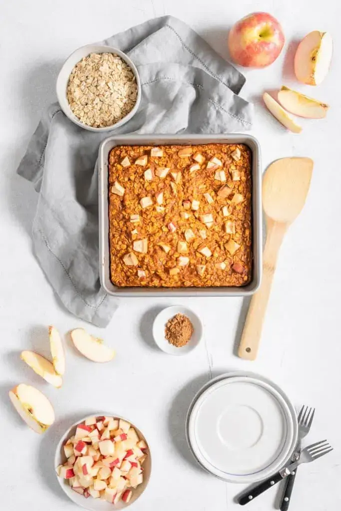 Baked apple and pumpkin oatmeal in a square pan on a table next to a wooden spoon.