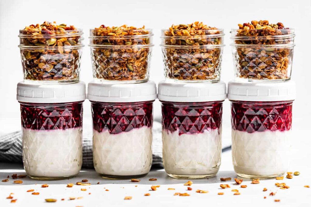 4 jars of Greek yogurt topped with a berry sauce with a separate container of granola on top. 