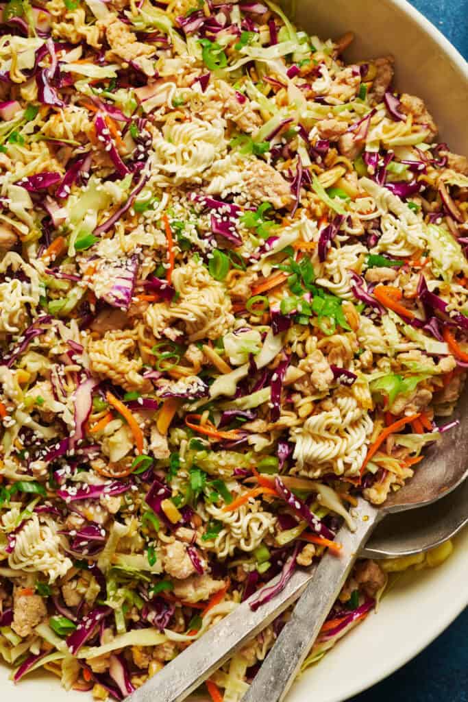 A close-up of chicken ramen salad in a white bowl with serving spoons.