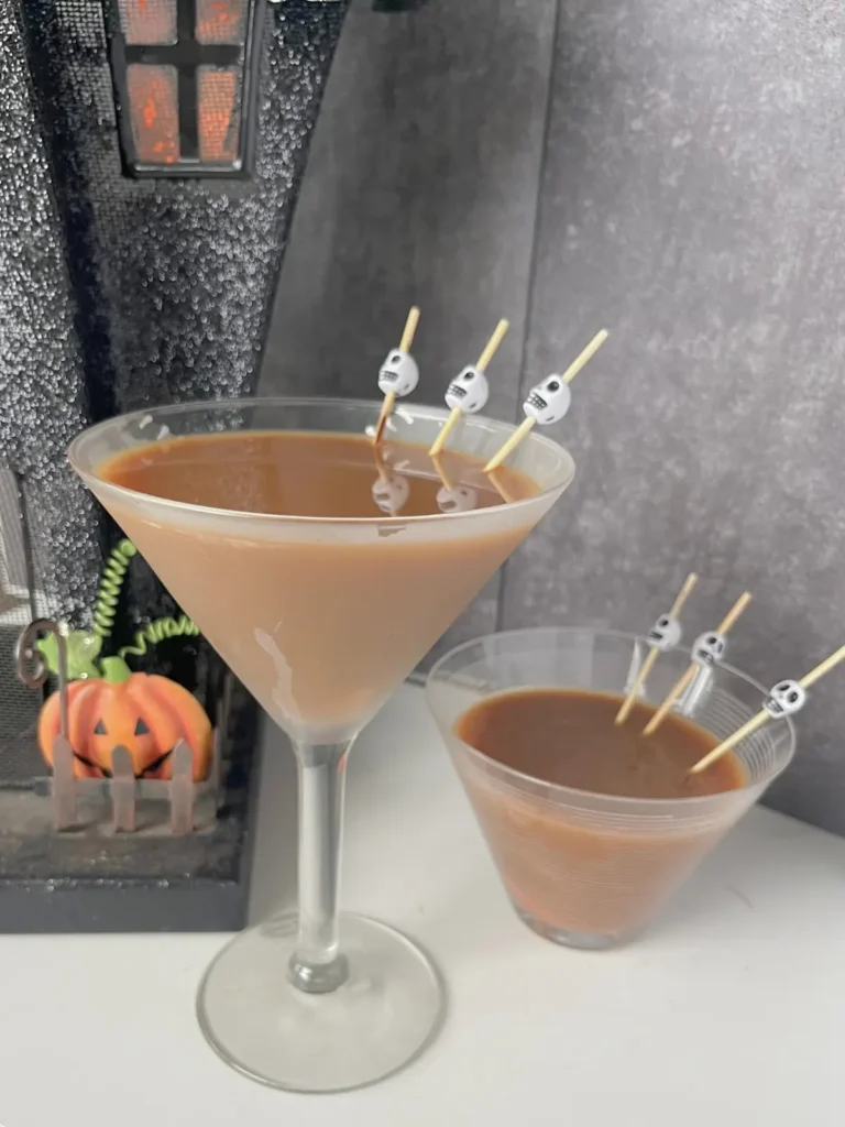 Two chocolate espresso martinis with wooden skull toothpicks in them next to a haunted house.