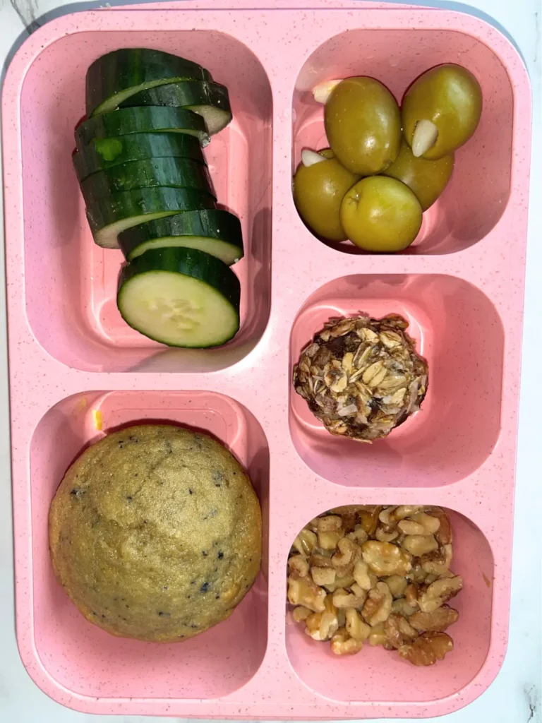 Garlic stuffed olives, sliced cucumbers, a muffin, an oatmeal ball, and chopped walnuts in a lunchable container.