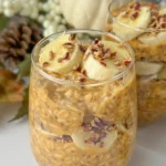 Two glasses full of pumpkin cheesecake protein overnight oats topped with banana slices and chopped pecans on a counter with a pumpkin decoration in the background.