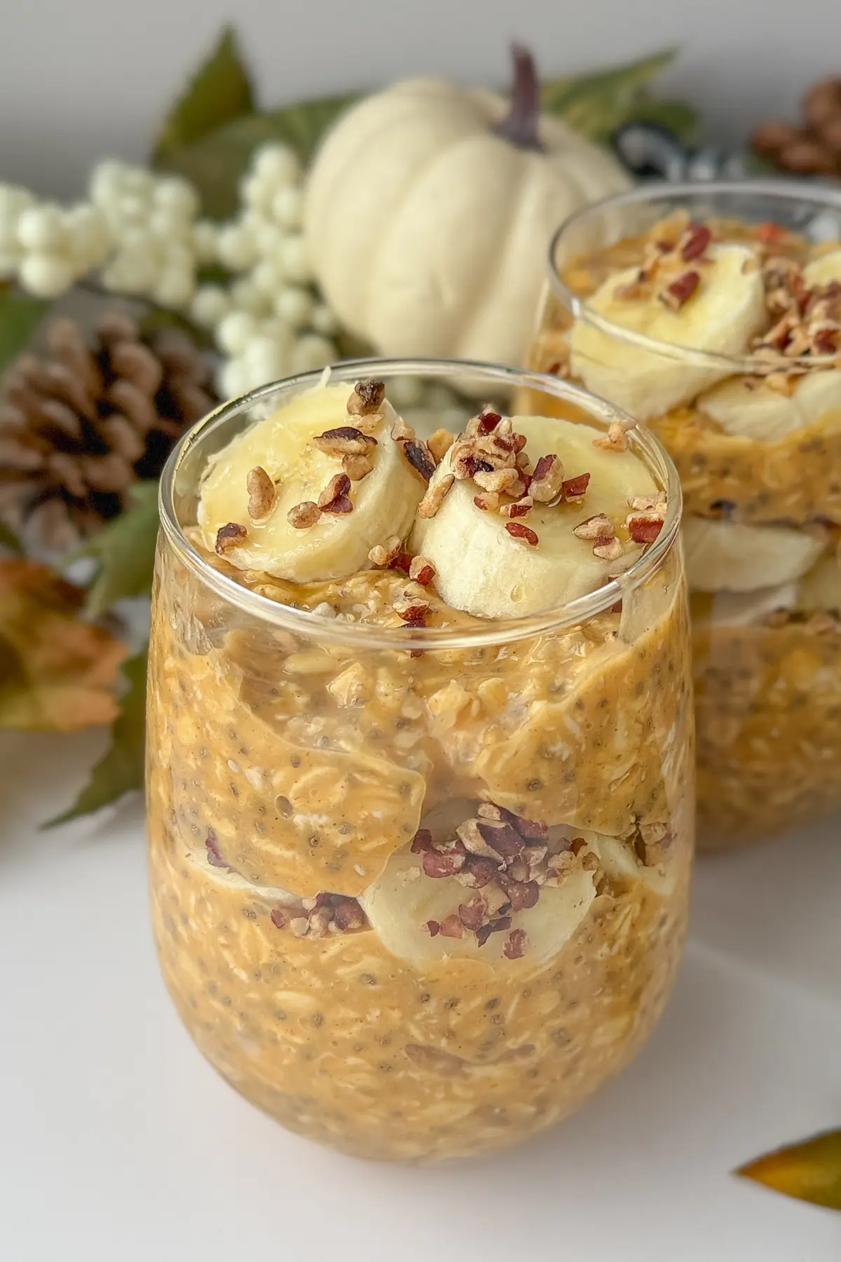 Two glasses full of pumpkin cheesecake protein overnight oats topped with banana slices and chopped pecans on a counter with a pumpkin decoration in the background.