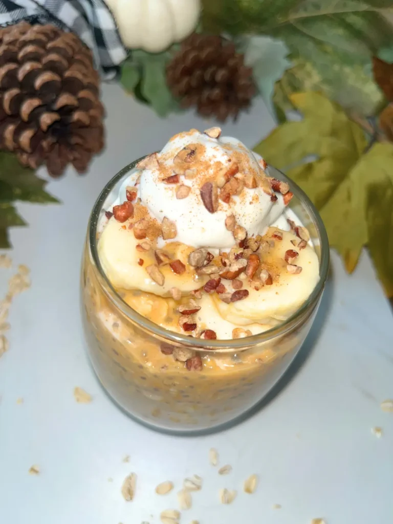 A close-up of pumpkin cheesecake overnight oats with protein powder in a glass jar, topped with whipped cream, chopped pecans and banana slices.