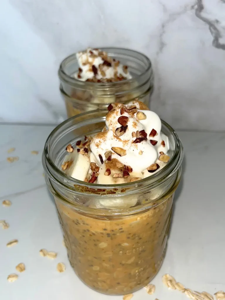 Two glass jars of pumpkin cheesecake overnight oats with protein powder on a counter, topped with whipped cream and crushed pecans.