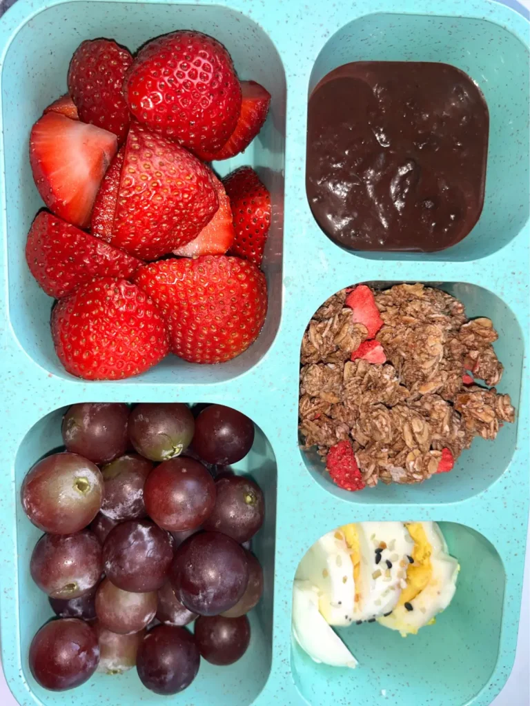 A snack box with diced strawberries, hard-boiled eggs, grapes, granola and a dark chocolate dipping sauce.