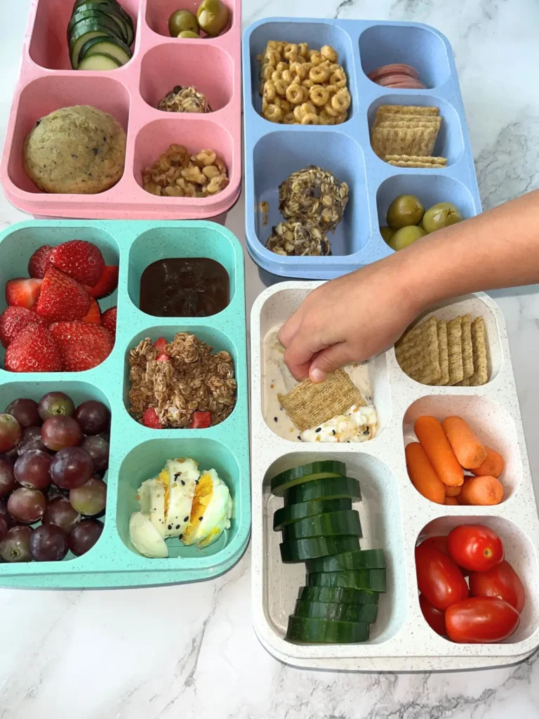 4 portable snack boxes filled with different variations of snacks on a counter, with a hand reaching into one of the compartments.