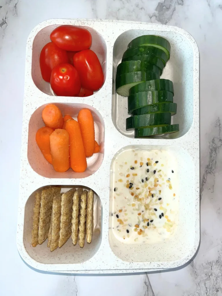 A portable lunchable container filled with cucumbers, tomatoes, carrots, whole wheat crackers and a Greek yogurt dip.