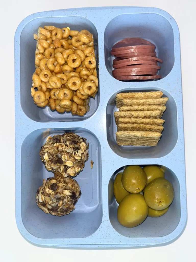 A snack box with 5 compartments containing cheerio peanut butter bars, oatmeal balls, salami, whole what crackers and olives.