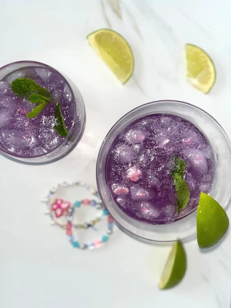 Two sparkly purple Enchanted cocktails on a counter next to lime wedges and friendship bracelets.