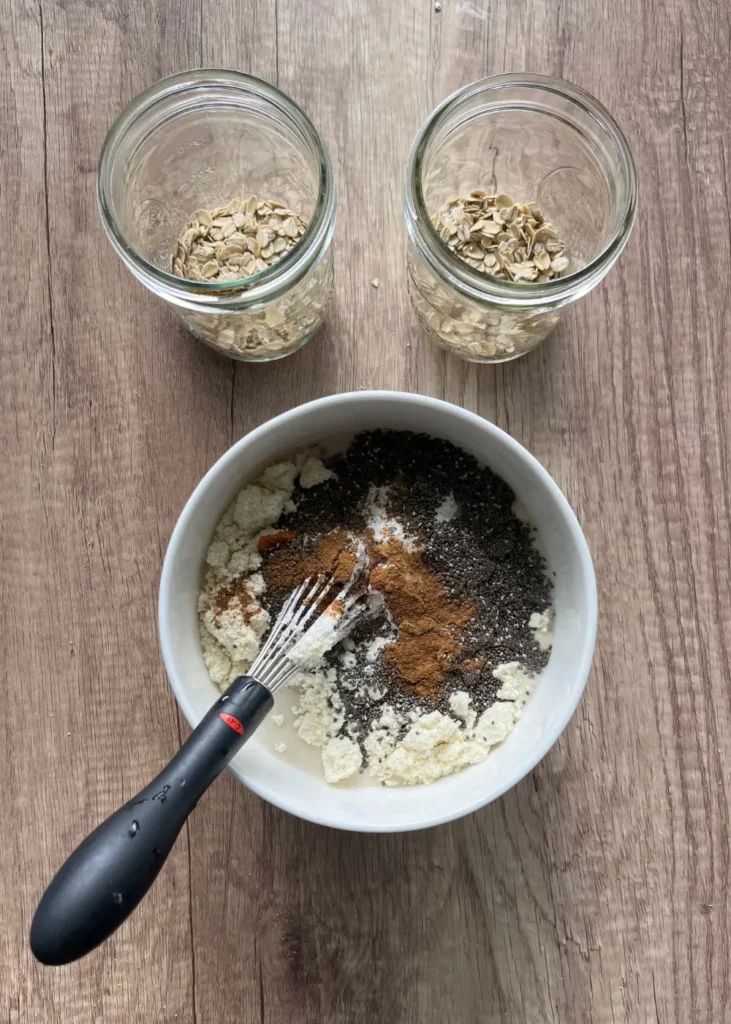 A bowl of the pumpkin cheesecake overnight oats mixture with a whisk in it next to two glass jars of oats.