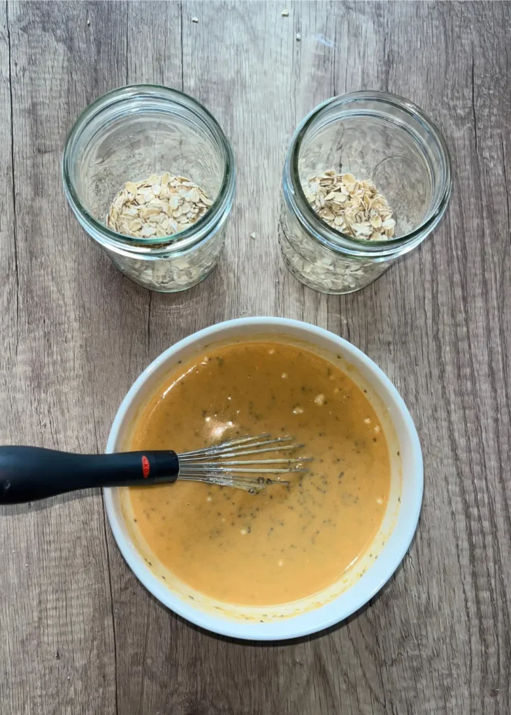 Two glass mason jars with rolled oats in it next to a bowl of the overnight oats mixture and a whisk on a counter.