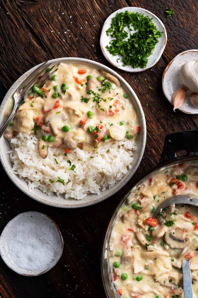 A bowl of creamy chicken a la king served over rice next to a pan of it on the table.