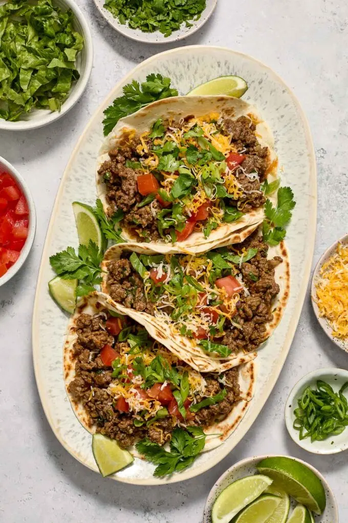 3 ground beef tacos on a platter topped with fresh herbs on a table with additional sides.