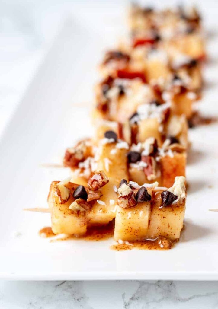 Skewers of caramel apples topped with walnuts and mini chocolate chips on a platter.