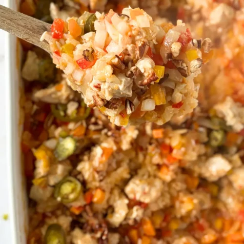 A big wooden spoonful of the coconut chicken and chopped pecan casserole over a casserole dish.