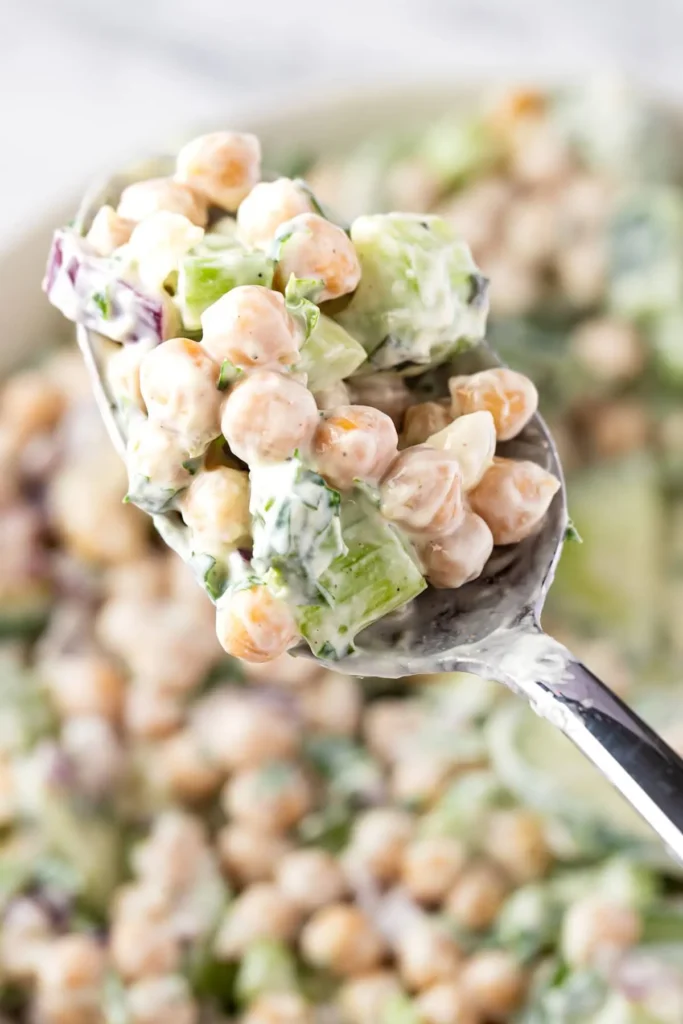 A close-up of chickpea salad with diced celery on a spoon with a bowl of it in the background.