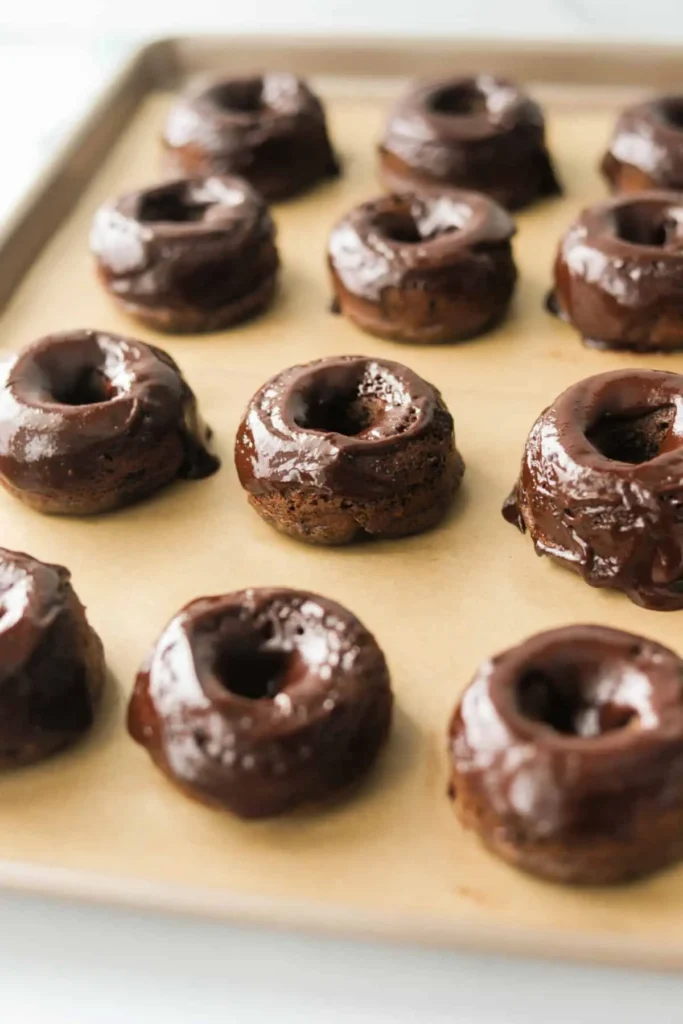 Baked and glazed chocolate donuts on a baking sheet lined with parchment paper. 